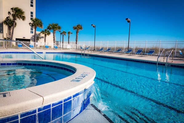 A swimming pool with a hot tub, lounge chairs, palm trees, and a white fence.