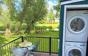 A balcony with a washing machine, a small table with chairs, and a view of a garden with trees and a pond. - WATER FRONT- Cozy Country Home SPA, Pristine Lawns, wildlife & trails to Explore (Spokane)