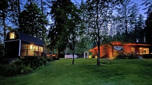 A rustic cabin with a wooden deck and a small porch, surrounded by a well-maintained lawn and tall trees. - WATER FRONT- Cozy Country Home SPA, Pristine Lawns, wildlife & trails to Explore (Spokane)