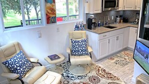 A modern kitchen with white cabinets, a stainless steel refrigerator, and a built-in microwave. There are two chairs with blue patterned cushions, a small table, and a large window with a view of trees. - WATER FRONT- Cozy Country Home SPA, Pristine Lawns, wildlife & trails to Explore (Spokane)