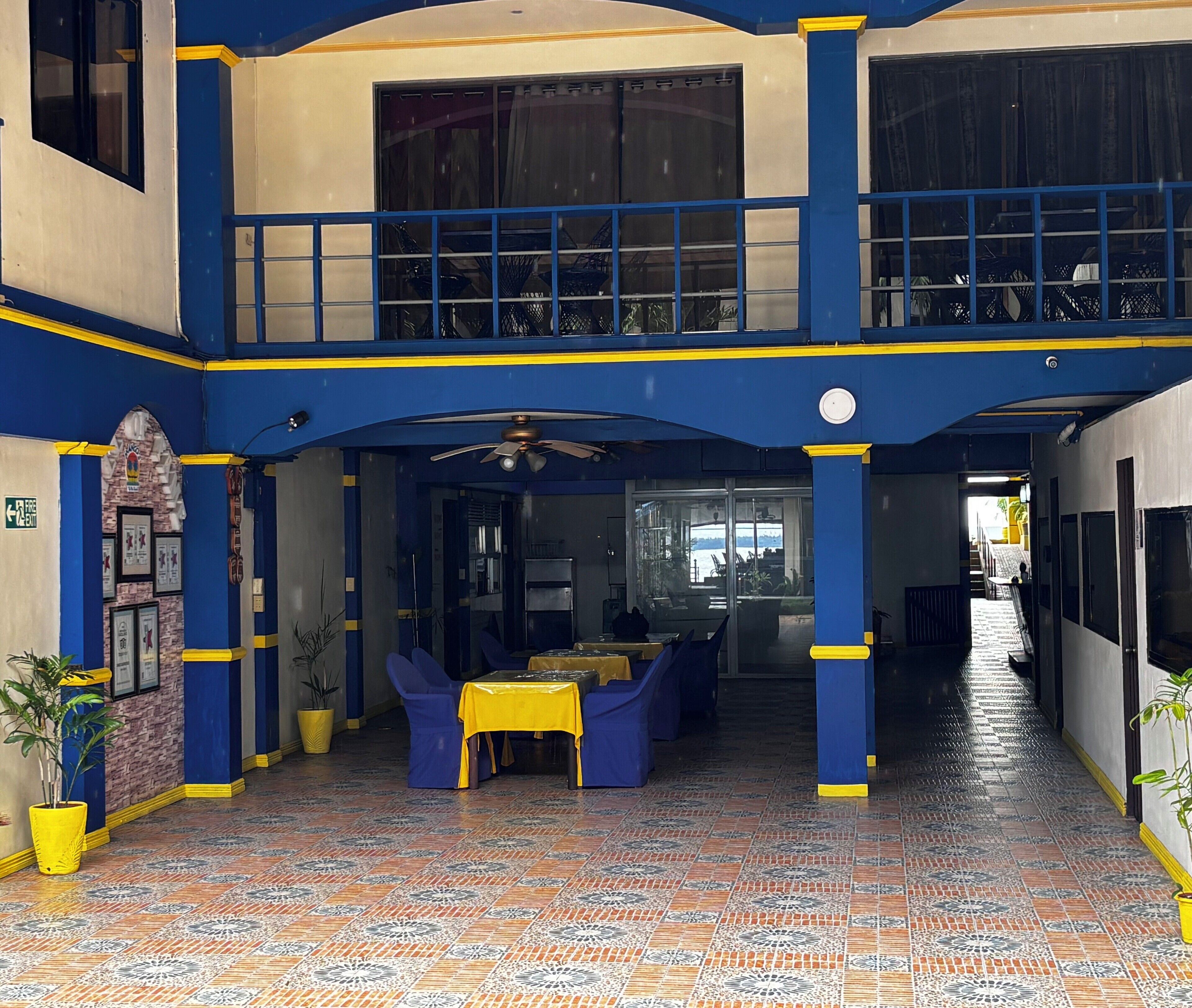 Un vestíbulo amplio con suelo de baldosas, decoración azul y amarilla, y una zona de estar con mesas y sillas.