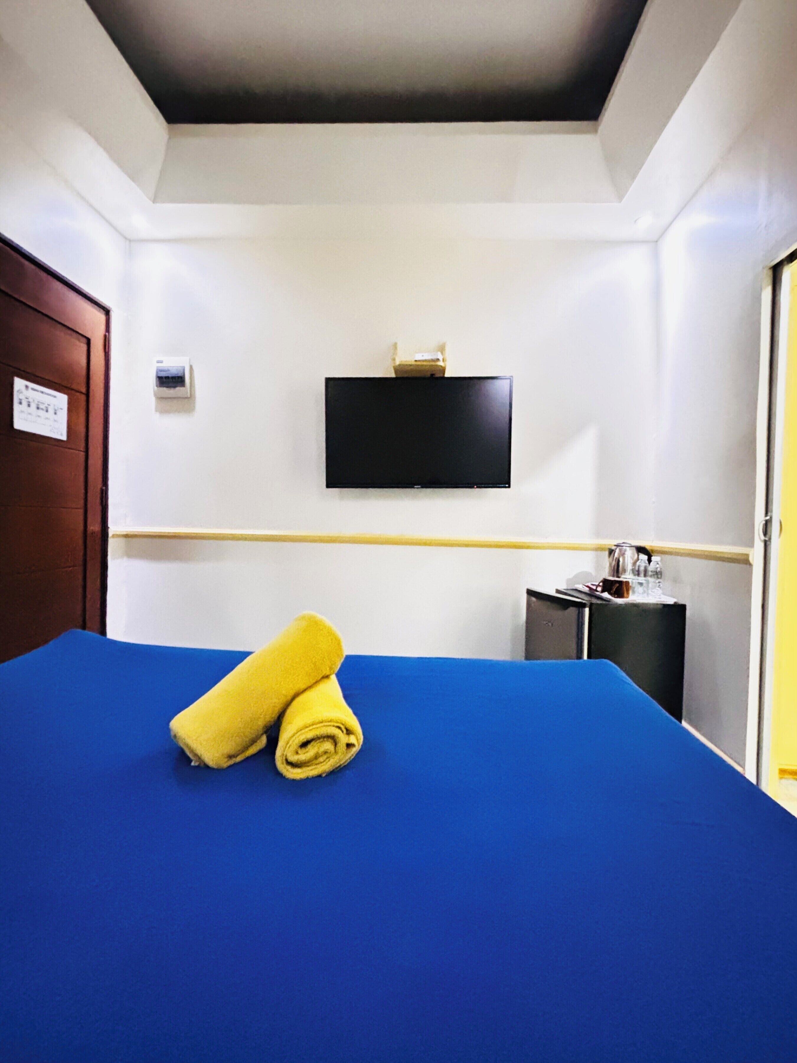 Una camera d'albergo con un letto blu, una televisione fissata al muro, una porta di legno e un piccolo mobile con oggetti sopra.