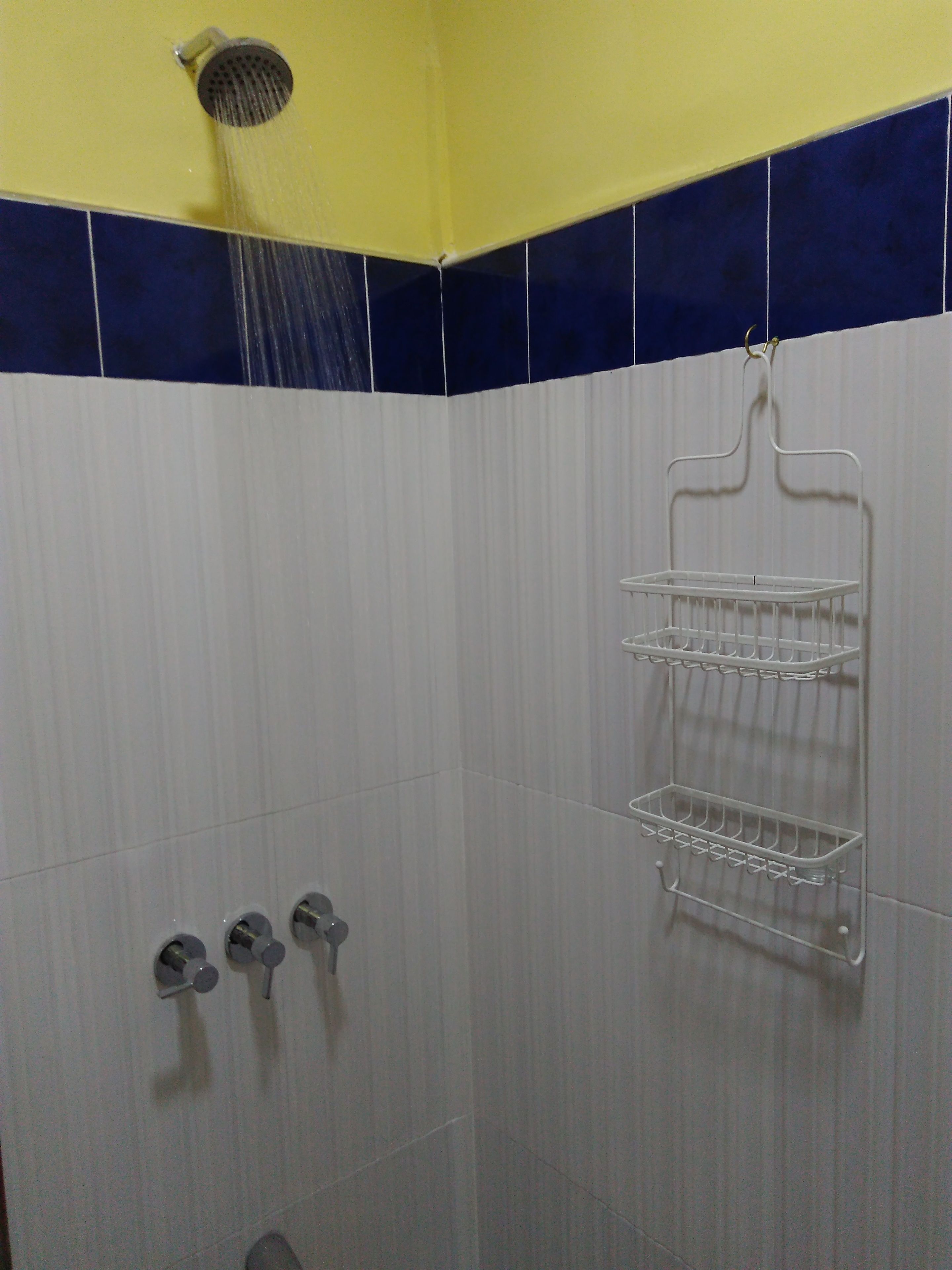 A shower area with a white tiled wall, a yellow ceiling, and a white shelf with three compartments.
