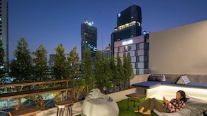 A rooftop terrace with a bean bag chair, a small table, and a sofa. There are trees and a view of city buildings in the background. - Natee Hostel (Bangkok)