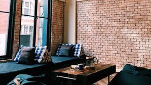 A cozy room with a brick wall, large windows, and a seating area with cushions and a coffee table. - Natee Hostel (Bangkok)