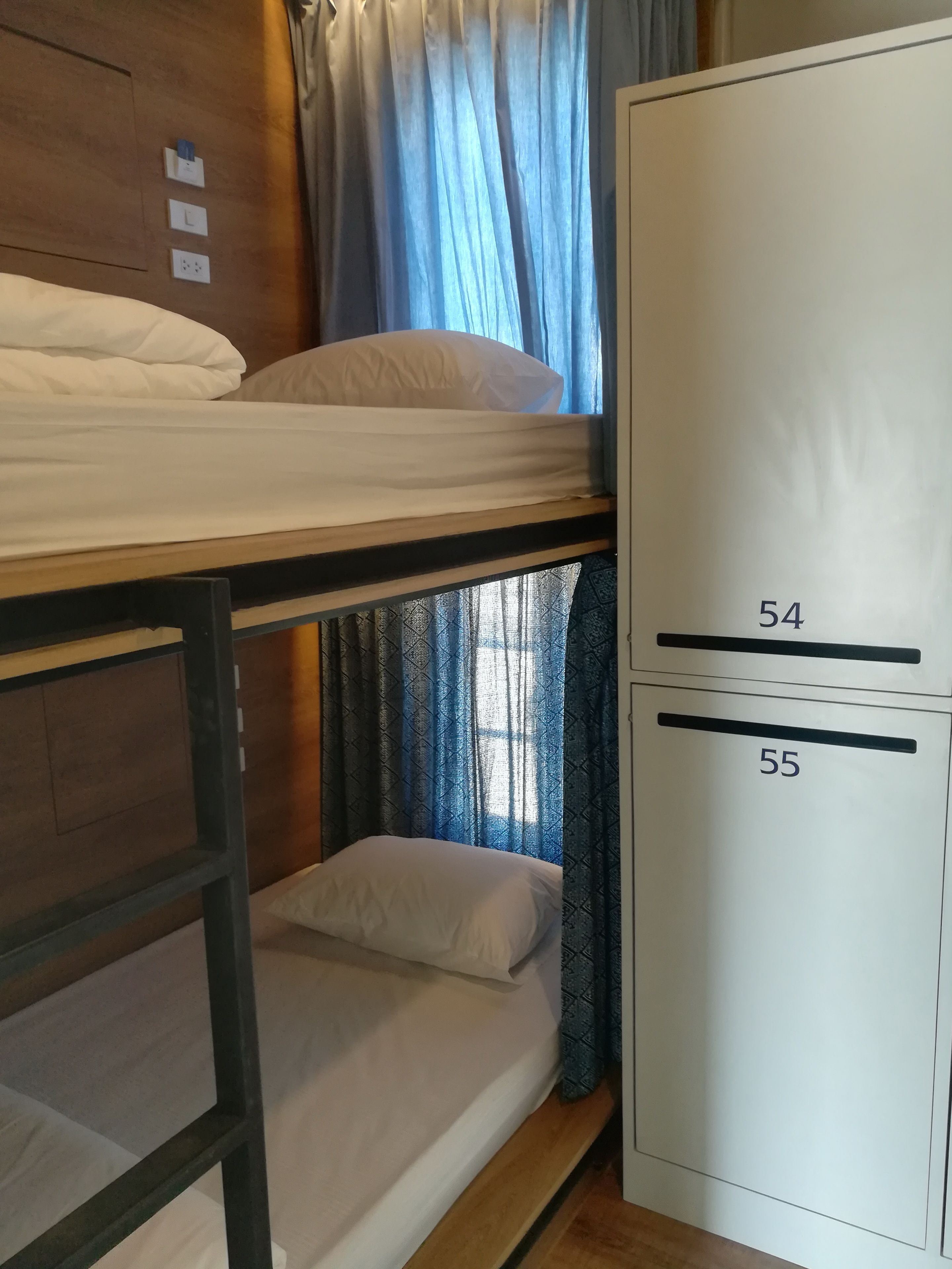 A bunk bed with two levels, a wardrobe with numbers 54 and 55, and a window with blue curtains.