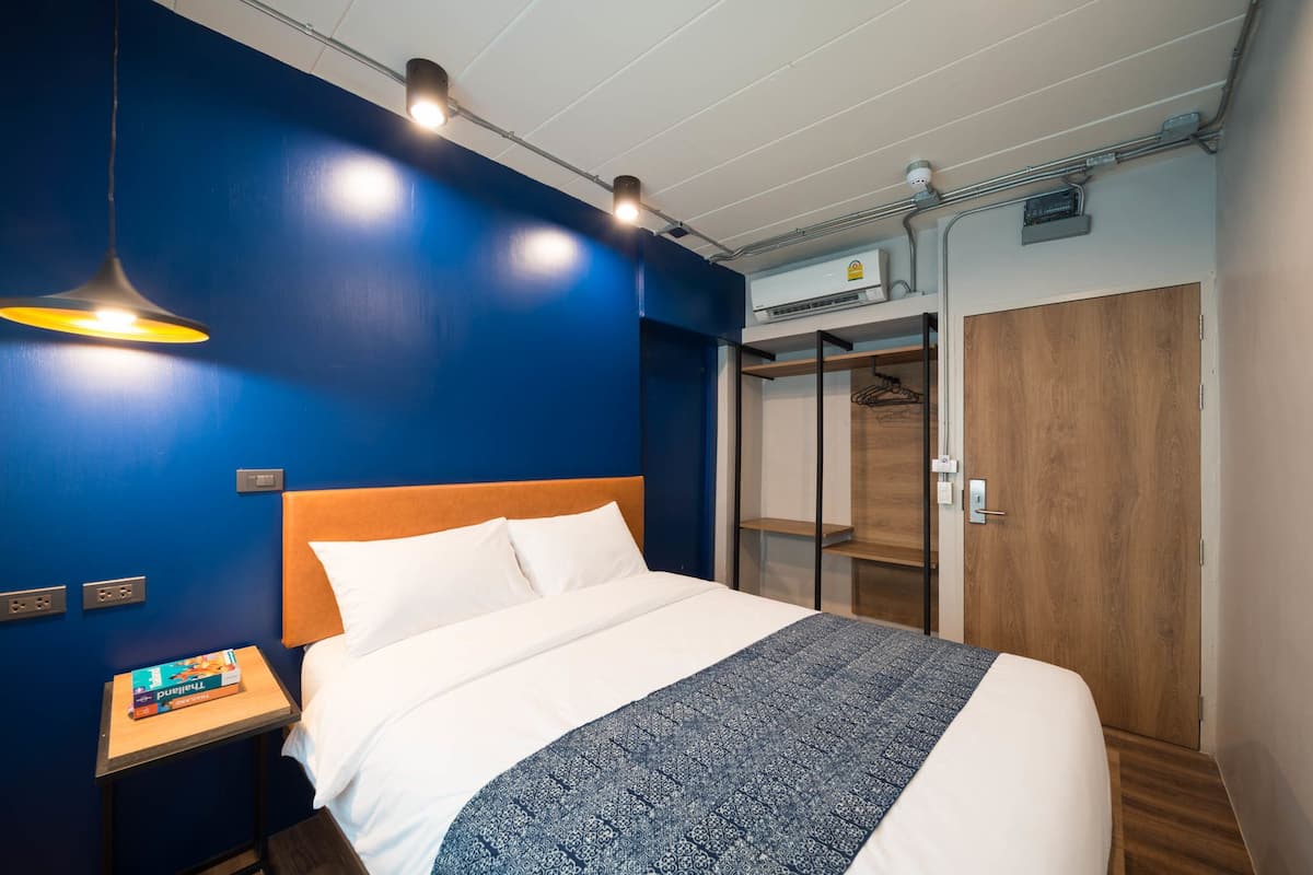 A modern hotel room with a large bed, a bedside table with a book, a wardrobe, and a hanging light fixture.