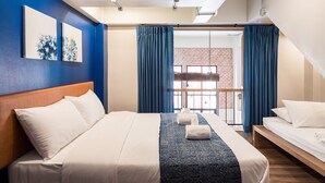 A hotel room with a large bed, a wooden headboard, a blue accent wall, and a view of a brick building through a glass door. - Natee Hostel (Bangkok)