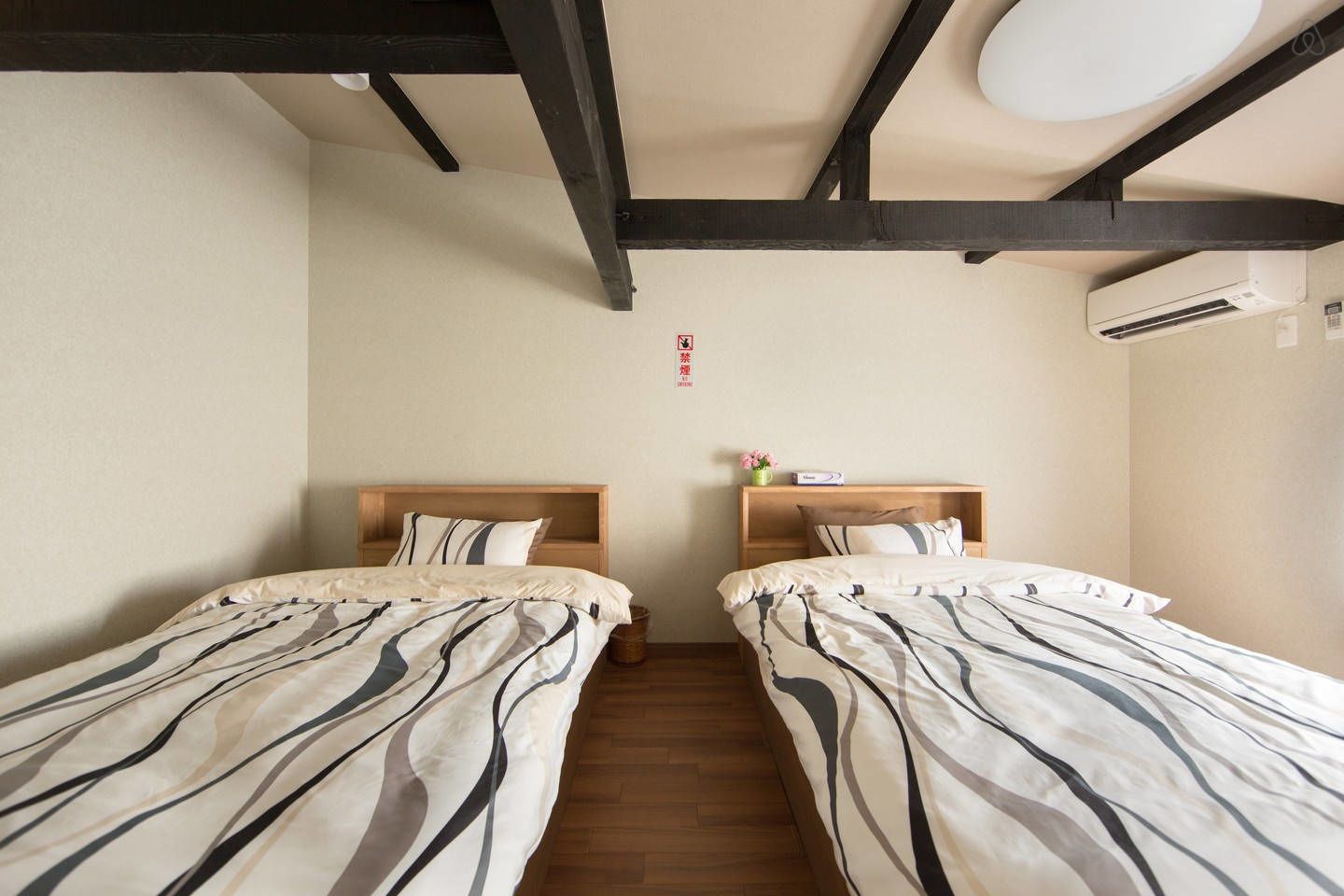 A hotel room with two beds, wooden headboards, a small bedside table with a vase, and an air conditioning unit on the wall.