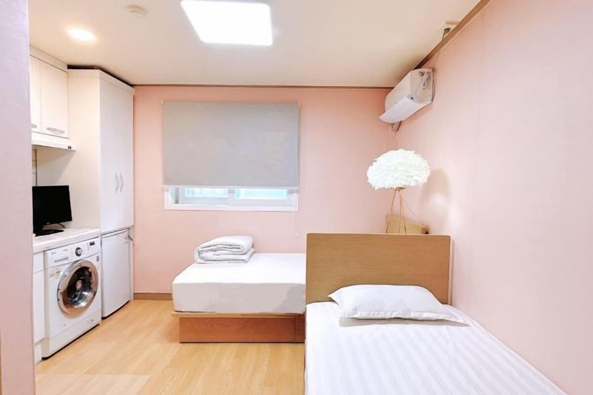 A compact bedroom with a single bed, a small bedside table, a washing machine, and a window with blinds.