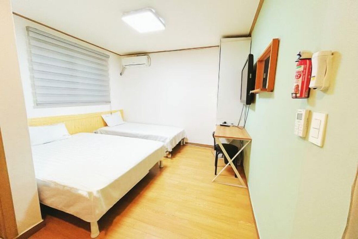 A small, well-lit room with two single beds, a desk, and a television.