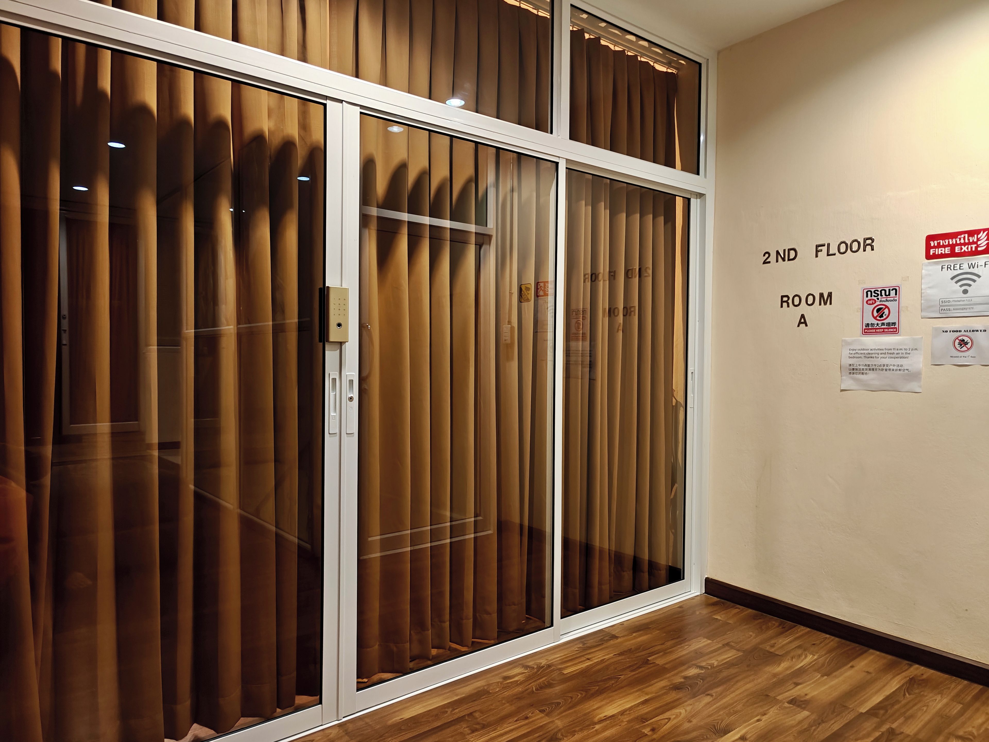 A hotel room with a sliding glass door, wooden blinds, a wooden floor, and a wall with room information and safety notices.