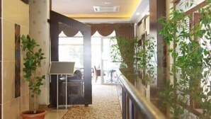 A hotel corridor with patterned carpet, a reception desk, potted plants, and a seating area in the background.