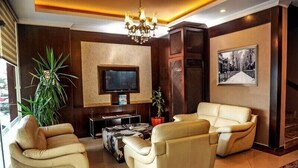 A modern living room with leather sofas, a flat-screen TV, a chandelier, and a large potted plant.