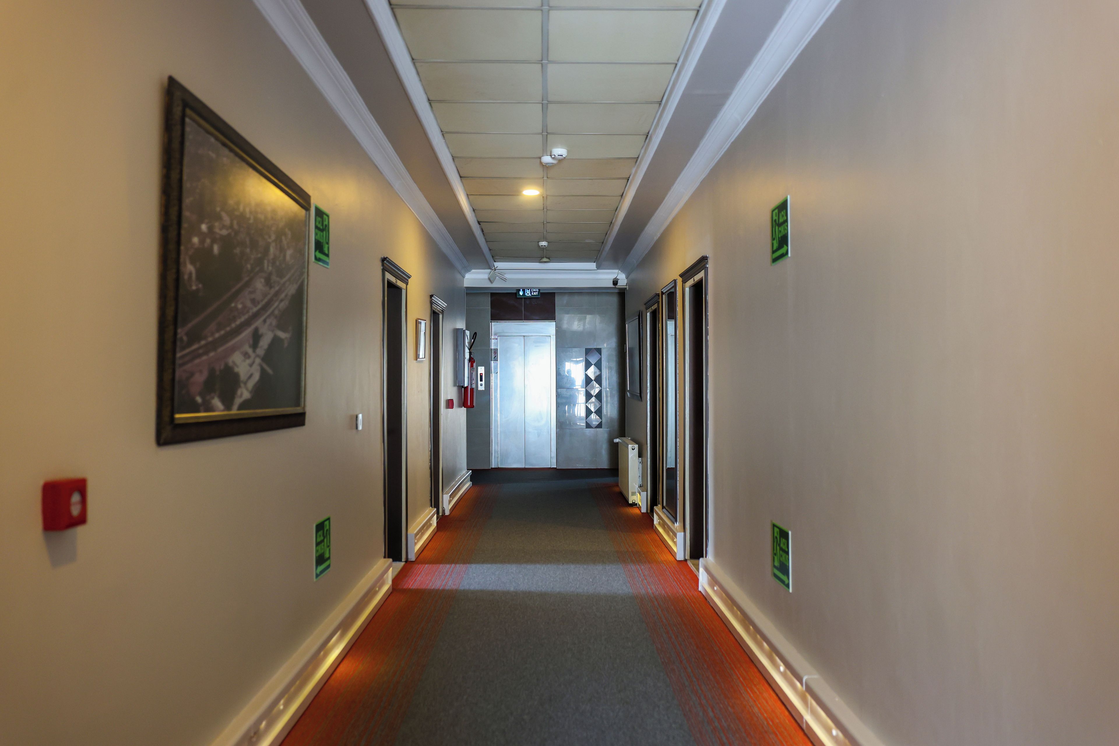 A corridor with doors on both sides, emergency exit signs, and a fire alarm on the wall.