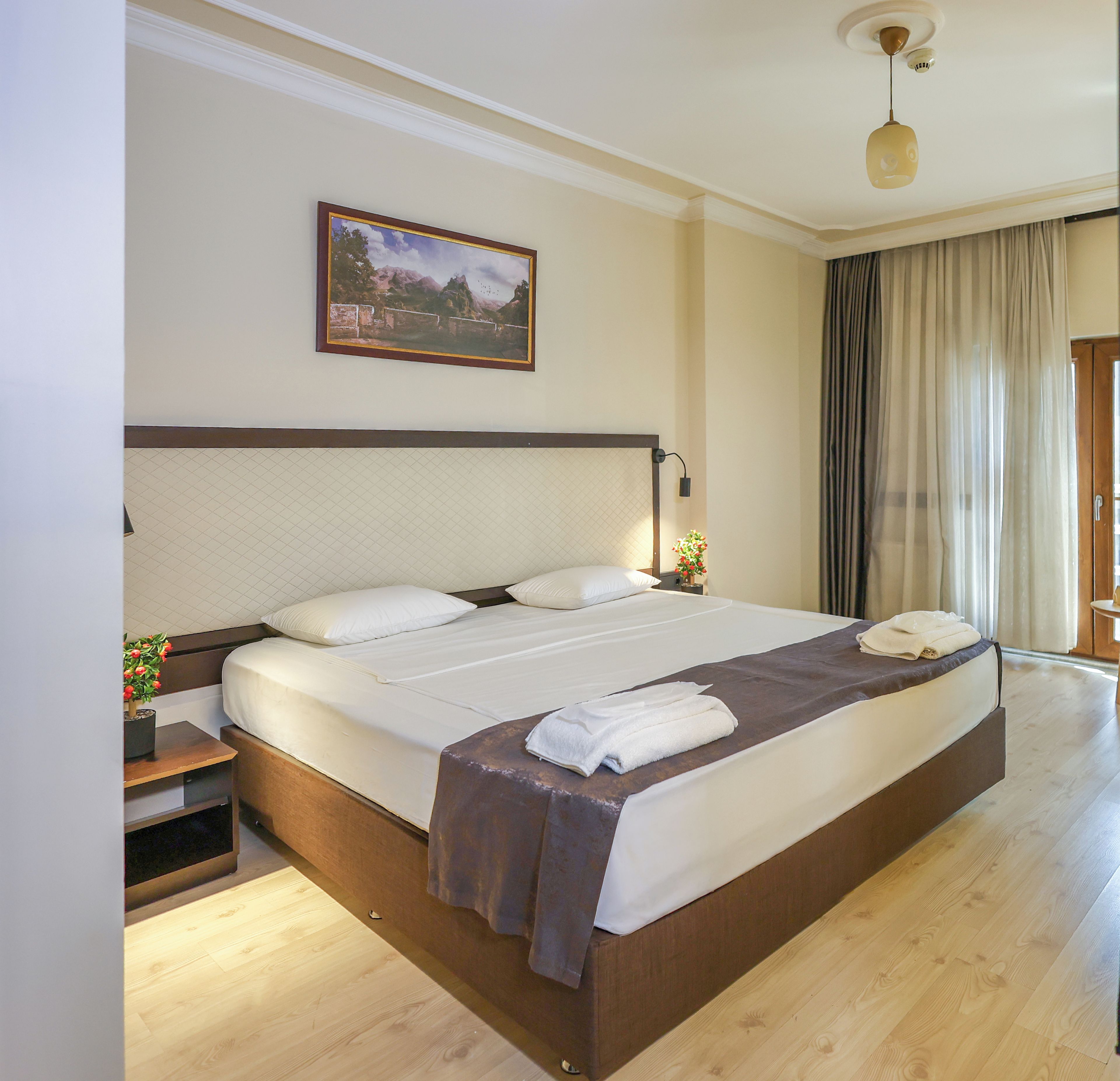 Una habitación de hotel con una cama grande, una mesita de noche, una lámpara, un cuadro en la pared y una ventana con cortinas.