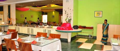 A restaurant with multiple dining tables and chairs, a counter with a red cloth, and a green wall with a framed picture.
