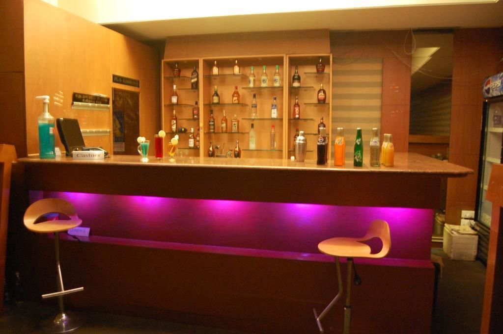Een bar met een toonbank, drankflessen op planken, twee barkrukken en een paars verlichte bar.