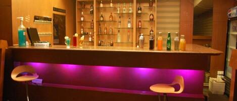 Een bar met een toonbank, drankflessen op planken, twee barkrukken en een paars verlichte bar.