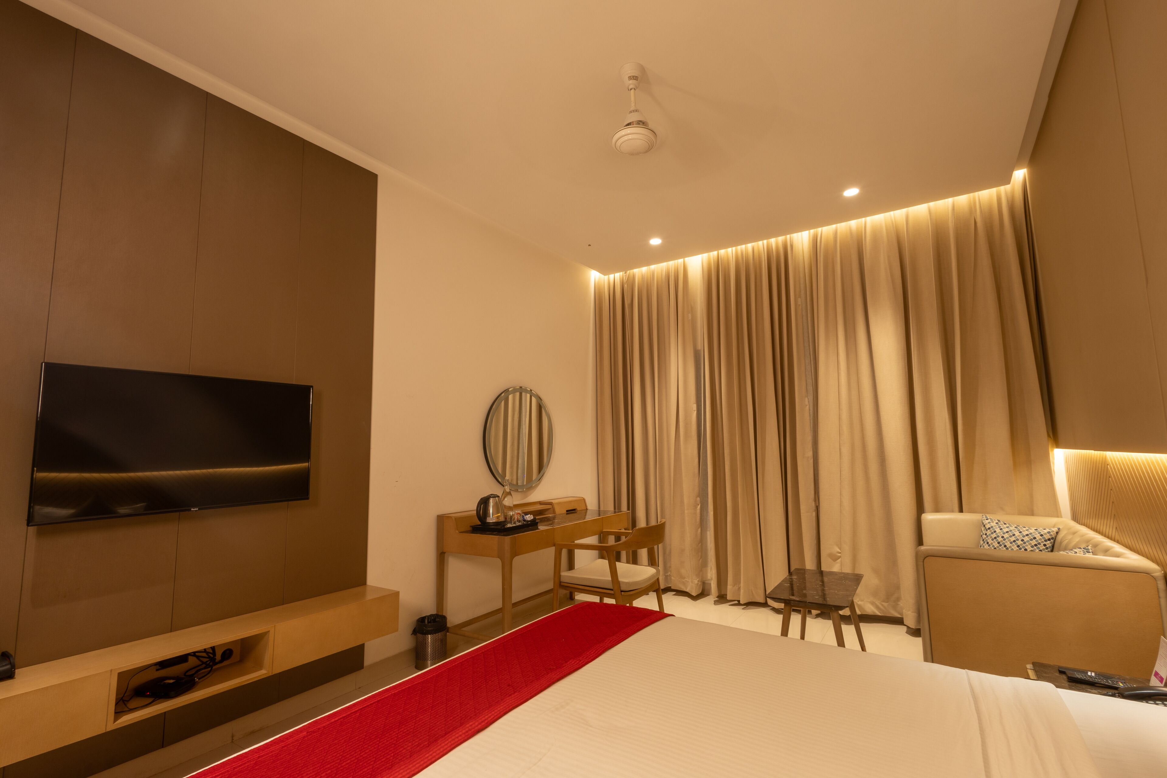 A modern hotel room with a large bed, a desk with a chair, a television, and a seating area with curtains.