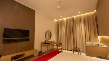A modern hotel room with a large bed, a desk with a chair, a television, and a seating area with curtains.