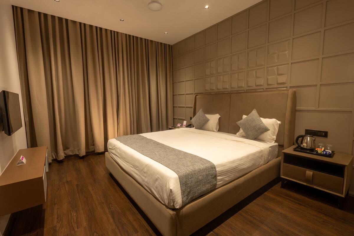 A modern hotel room with a large bed, a bedside table, a nightstand, a TV, and a large window with curtains.