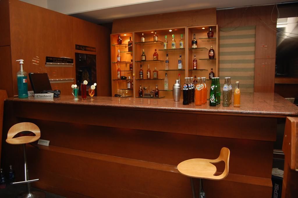 Una zona de bar con mostrador de madera, estanterías con varias botellas y dos taburetes de madera.