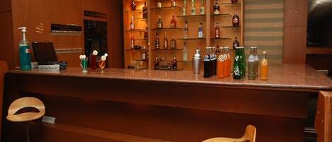 Una zona de bar con mostrador de madera, estanterías con varias botellas y dos taburetes de madera.