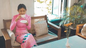 A woman in a pink dress sitting on a wicker sofa with a cushion, holding a white cup and saucer. There is a potted plant with yellow flowers on a table beside her. - 501 Merchant Bed & Breakfast - Hostel (Yangon)