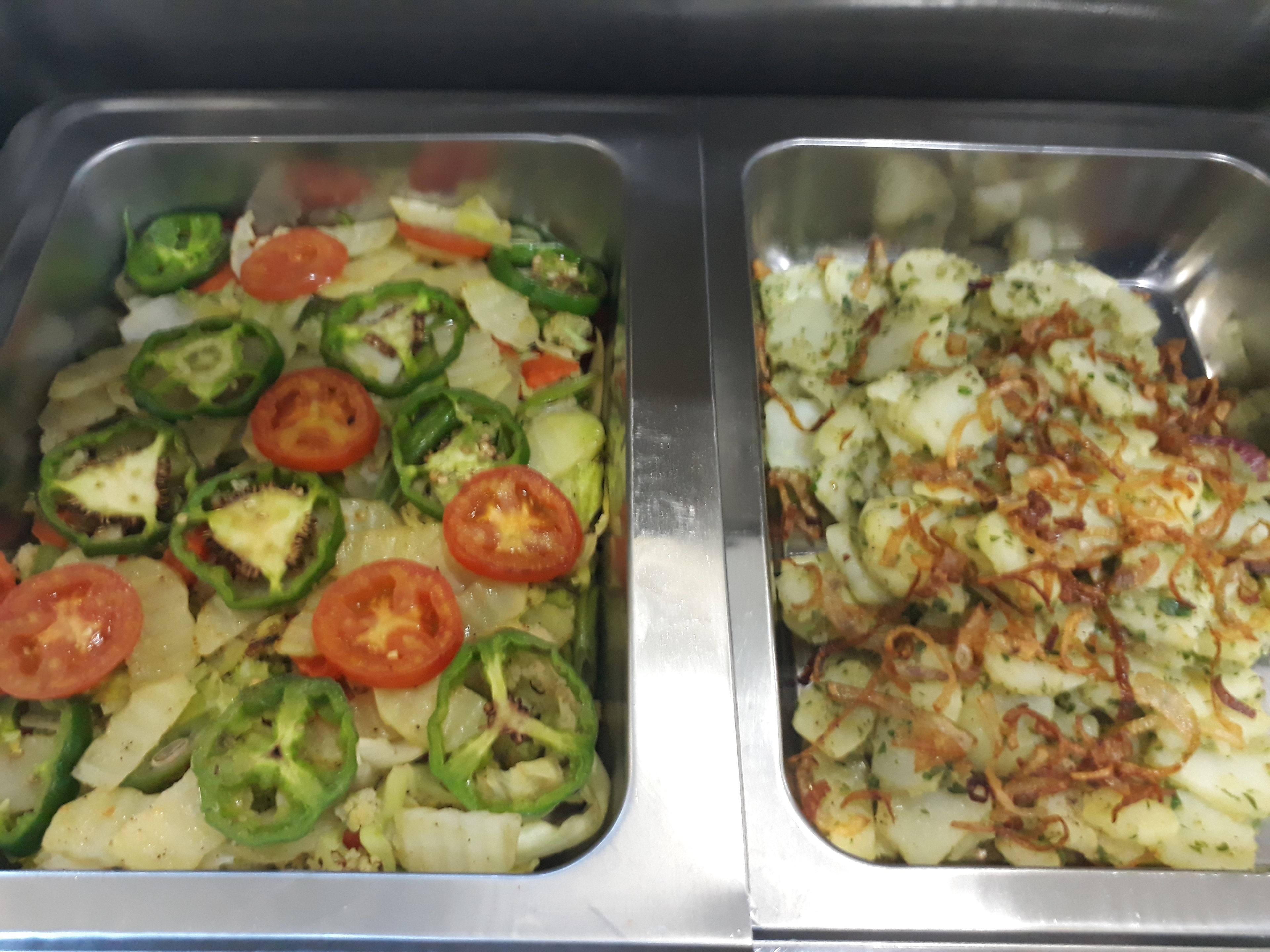 Dos bandejas de acero inoxidable con comida. Una tiene tomates en rodajas y pimientos verdes, y la otra, patatas con cebollas.