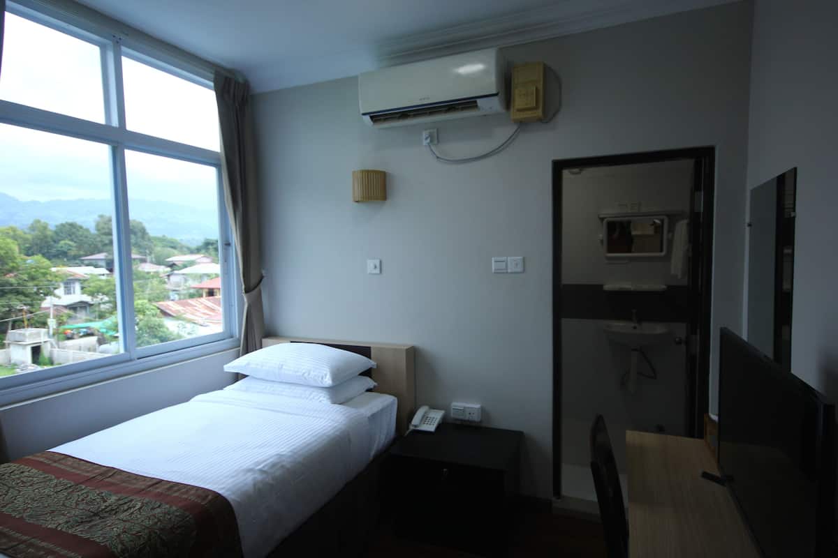 A hotel room with a bed, a bedside table with a phone, an air conditioner, a mirror, and a view of the outside.