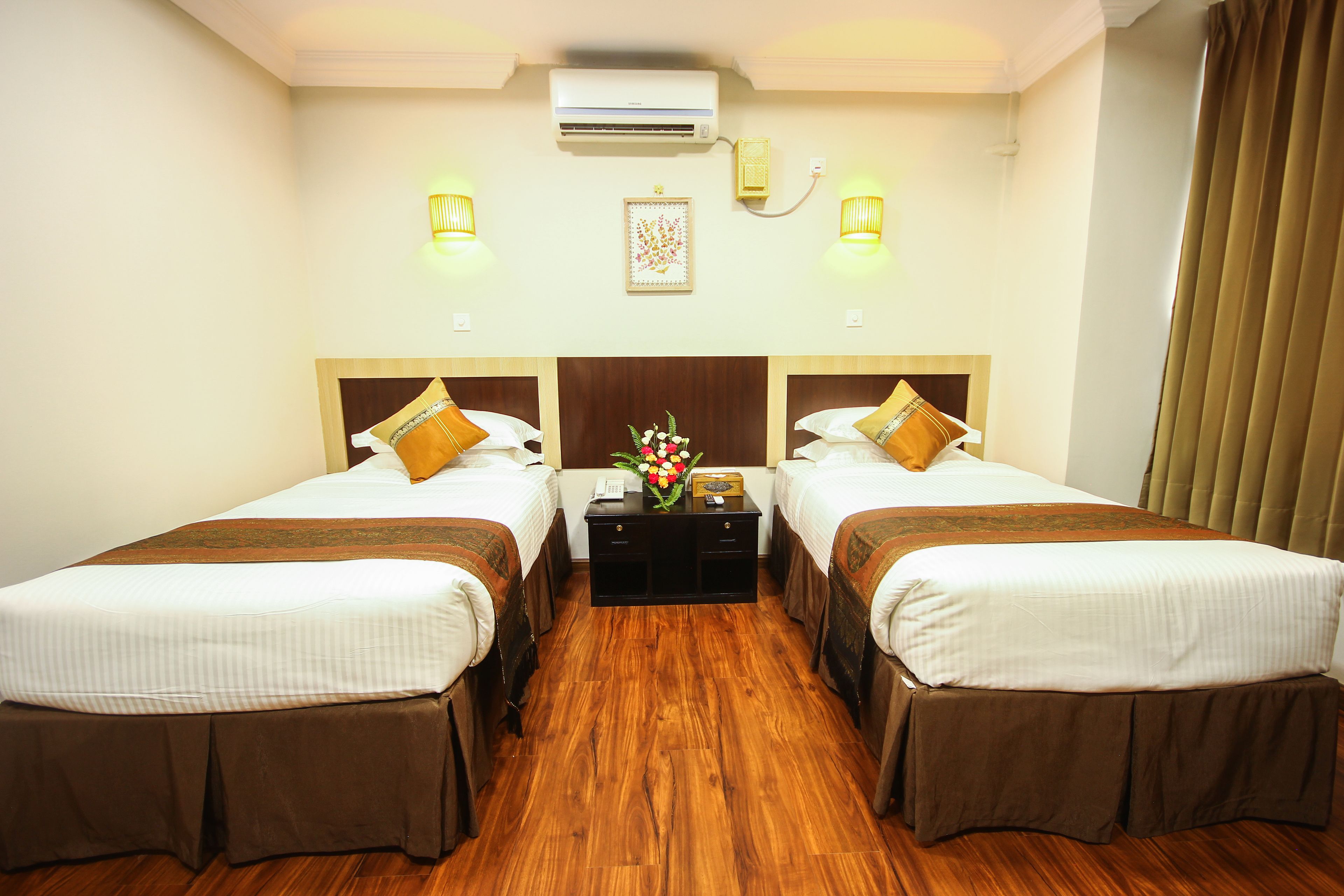 A hotel room with two single beds, a wooden floor, a nightstand with a vase of flowers, and wall-mounted lights.