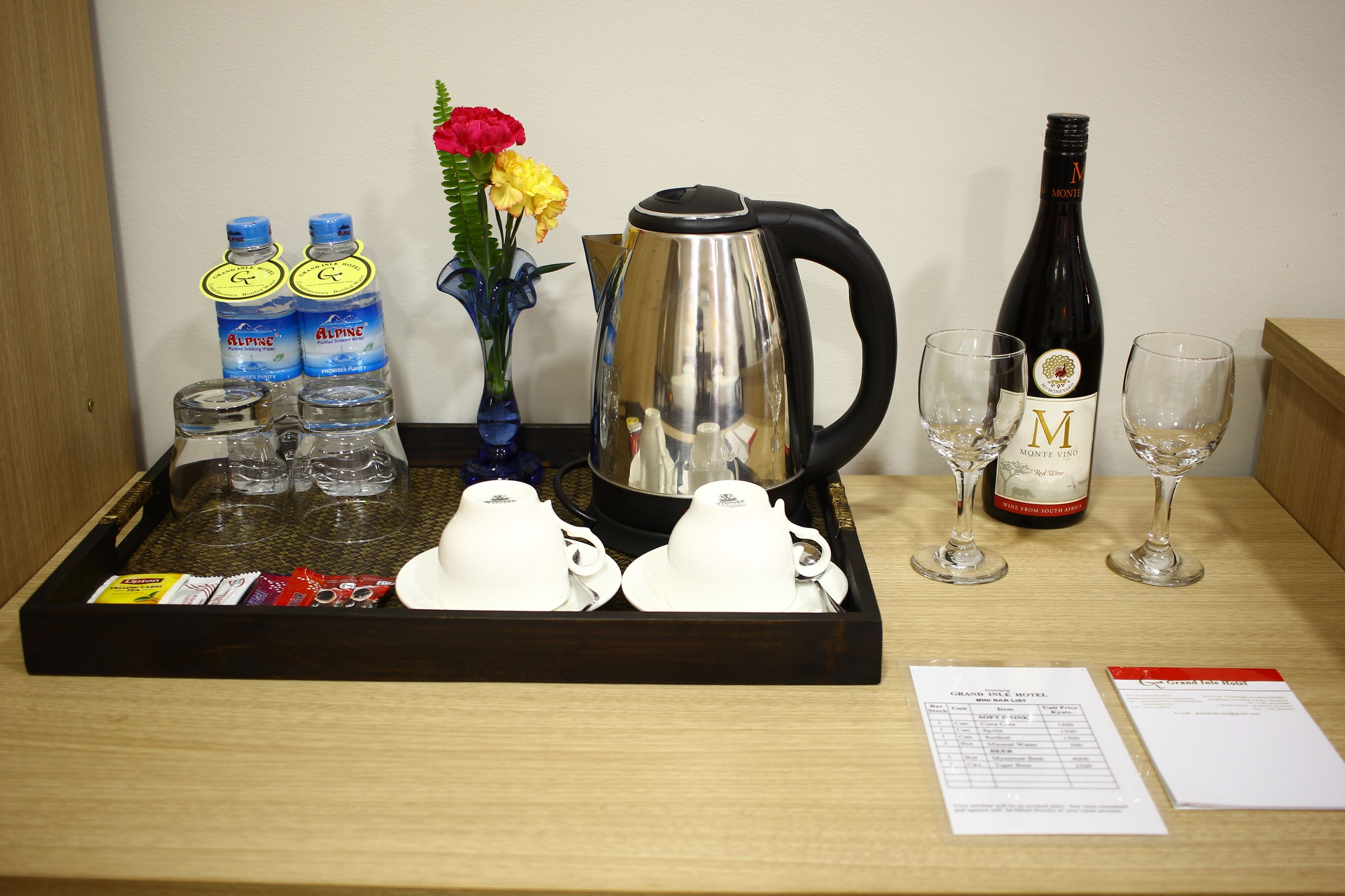 Una bandeja con botellas de agua, una cafetera, tazas de té, copas de vino y un menú sobre una mesa de madera.