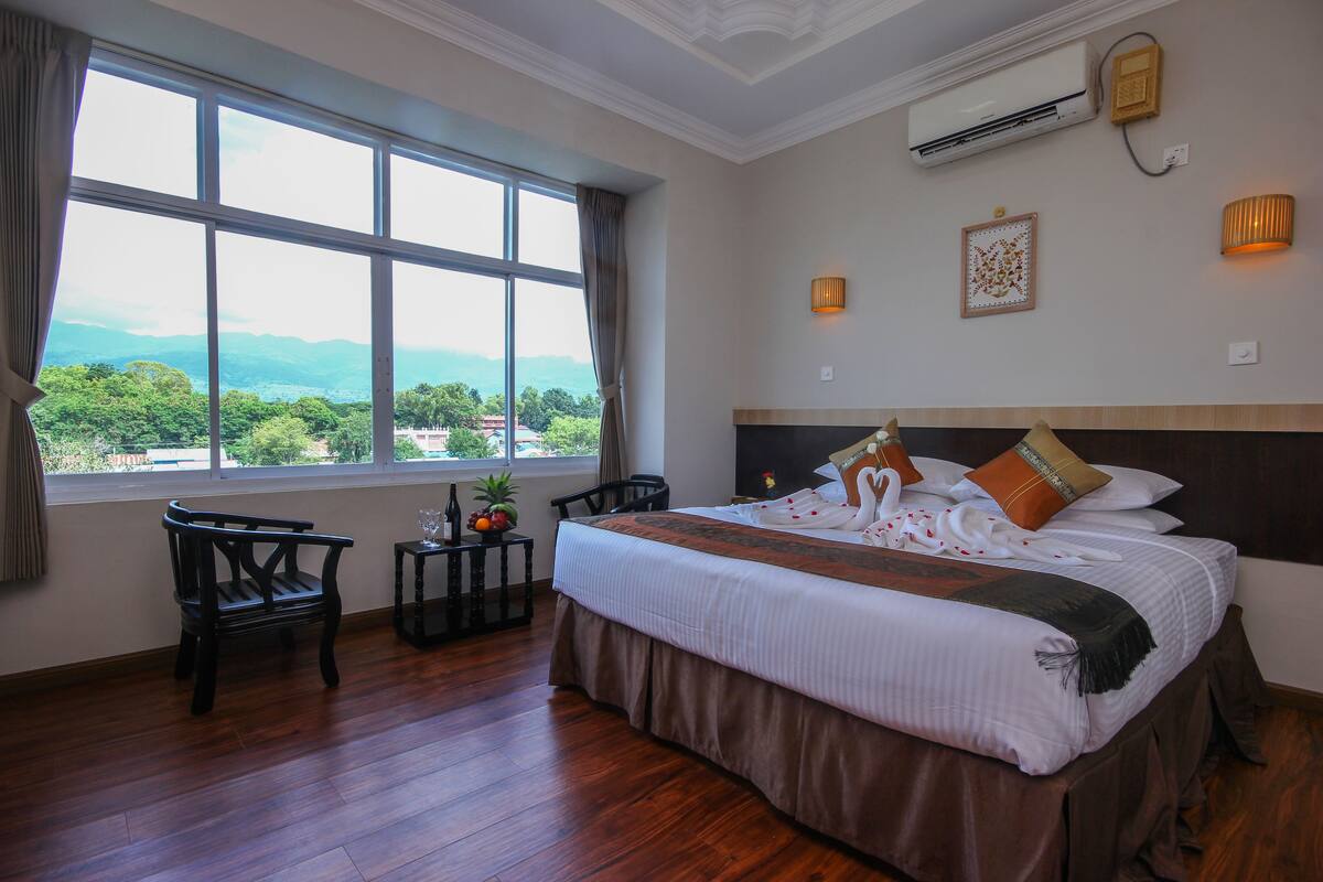 A hotel room with a large bed, two chairs, a small table with fruit, and a view of mountains through large windows.