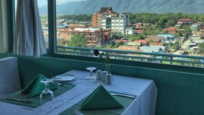 A dining table set with green napkins, plates, and cutlery, with a view of a town and mountains through a window. - Nyaung Shwe City Hotel (Nyaungshwe)