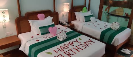 A hotel room with two beds, each with a green and white striped bedspread, heart-shaped pillows, and a 'Happy Birthday' message.