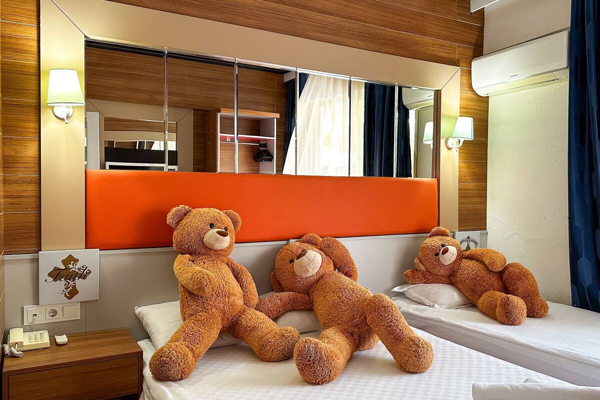 A hotel room with a large bed, a bedside table with a lamp, a mirror, and three teddy bears on the bed.