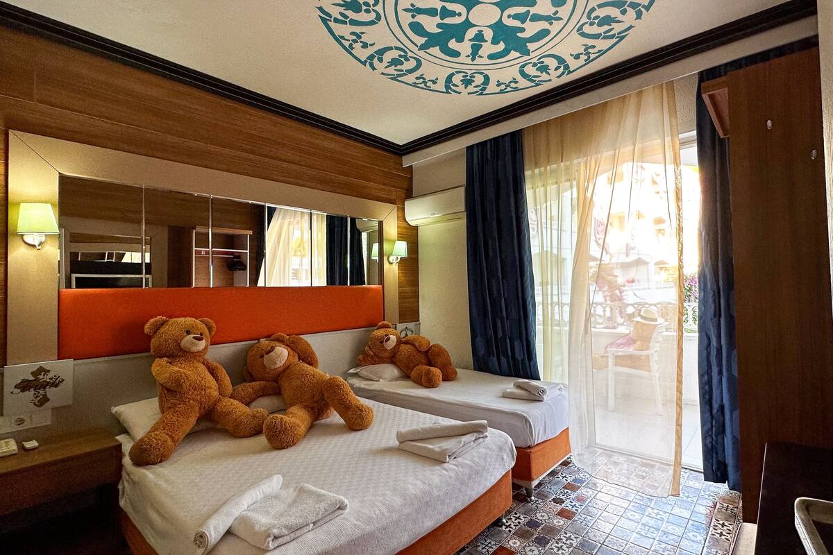 A hotel room with two beds, a large mirror, a teddy bear on one bed, and a view of the outside through a window.