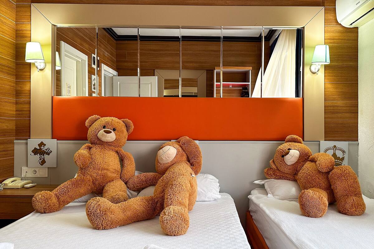 A hotel room with two beds, each with a teddy bear. There are wall-mounted lamps, a telephone, and a small table with a lamp.