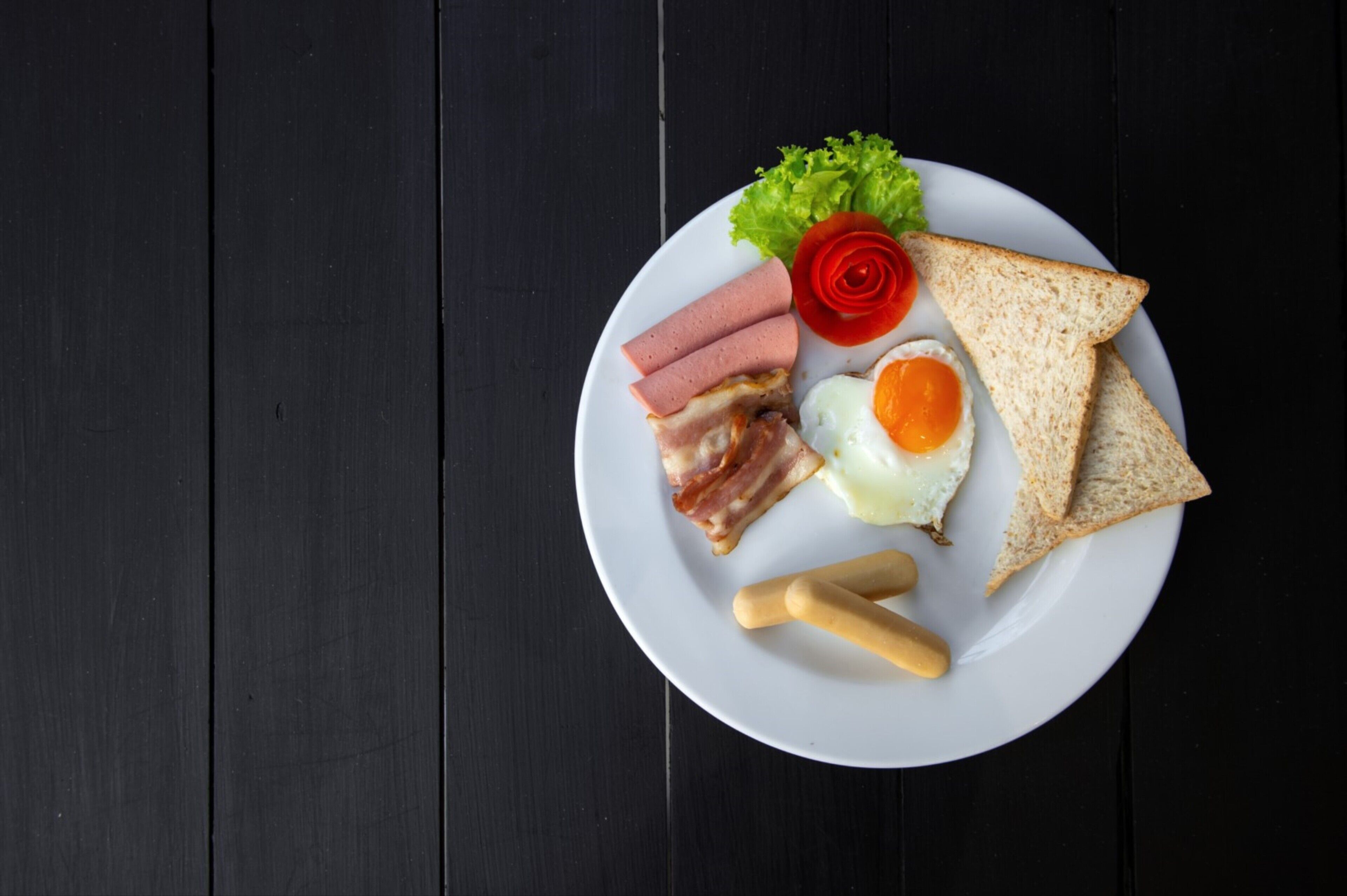 Une assiette blanche, posée sur une table en bois foncé, contient des tranches de bacon, des œufs, des toasts et une tomate.