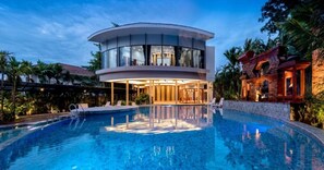 Une villa moderne dotée d’une structure circulaire en verre, d’une piscine et d’un bâtiment en pierre surmonté d’un toit rouge.