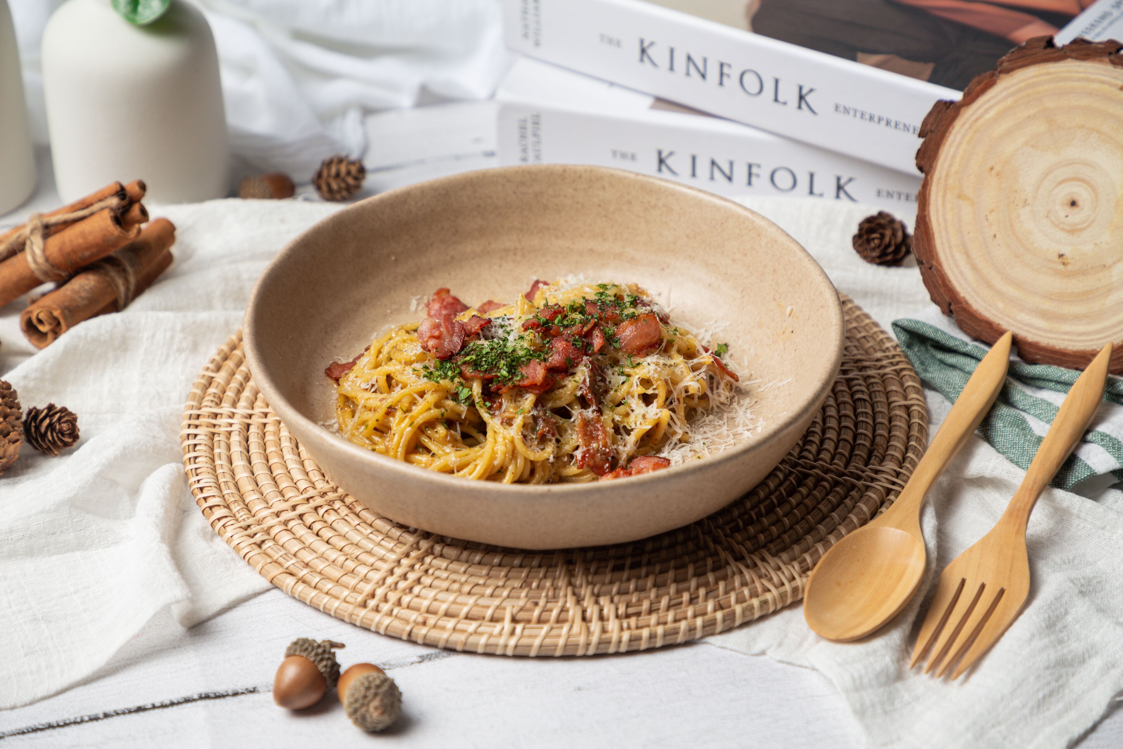 A bowl of pasta with cheese and herbs on a woven mat, surrounded by books, wooden utensils, and pine cones.