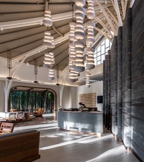 Un hall d’entrée spacieux avec un plafond haut, un éclairage moderne suspendu, un comptoir d’accueil et une vue sur des bambous à l’extérieur.
