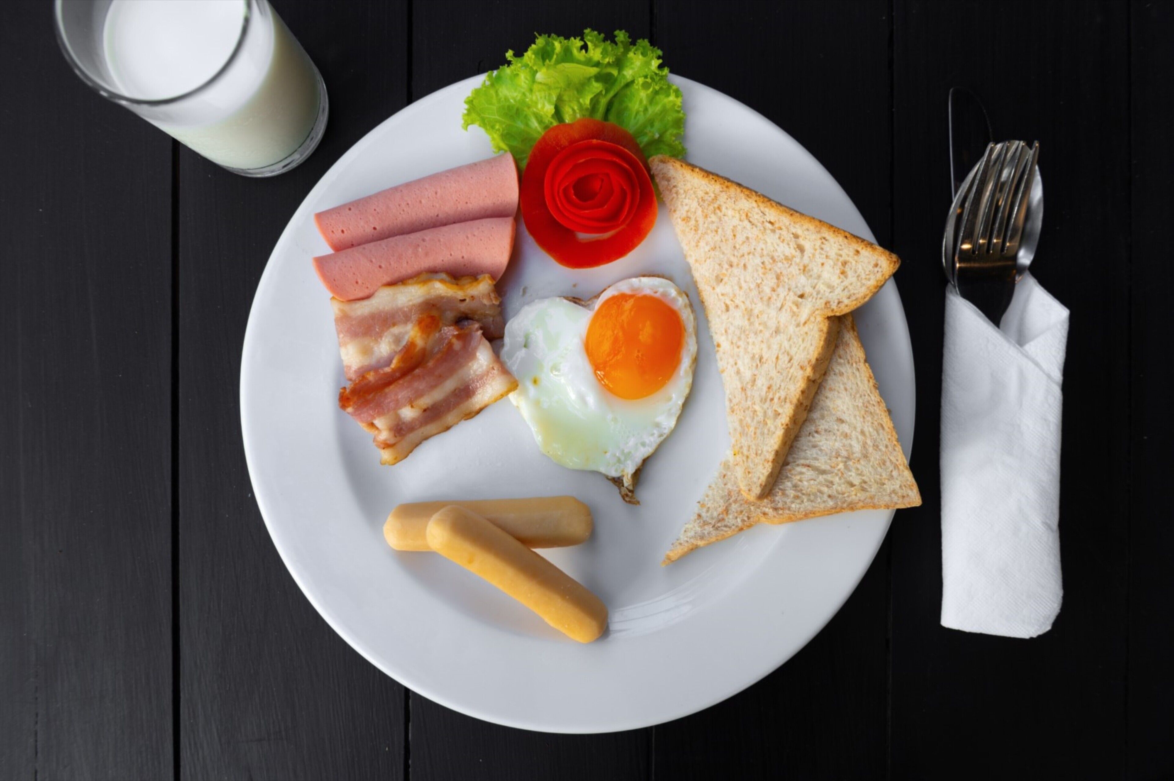 A plate with bacon, eggs, toast, and a glass of milk on a dark wooden table.
