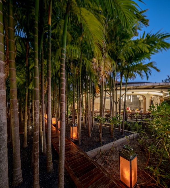 Un jardin intérieur paisible, agrémenté de grands plants de bambou, de passerelles en bois et de lanternes illuminées, qui mène à un espace de restauration avec des tables et des chaises.