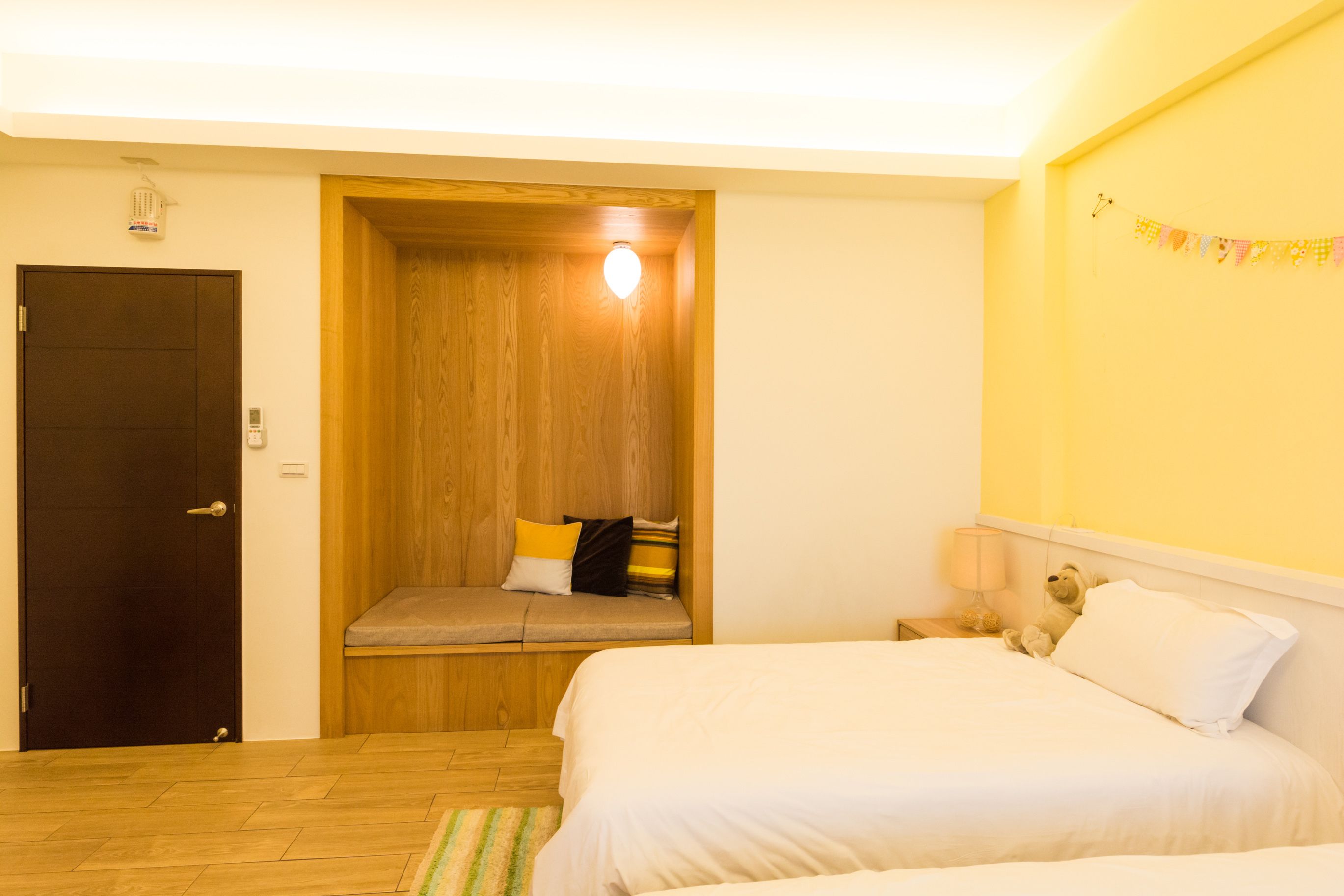 A bedroom with a wooden door, a bench with cushions, a bed with white bedding, and a nightstand with a lamp.