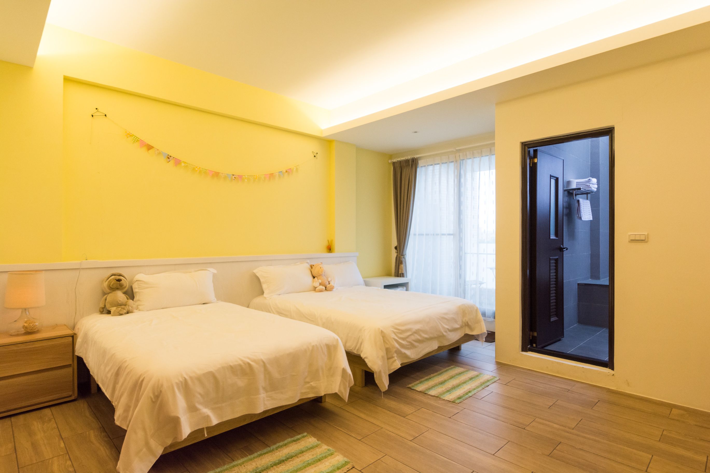 A hotel room with two beds, a small bedside table with a lamp, a teddy bear, and a curtain.