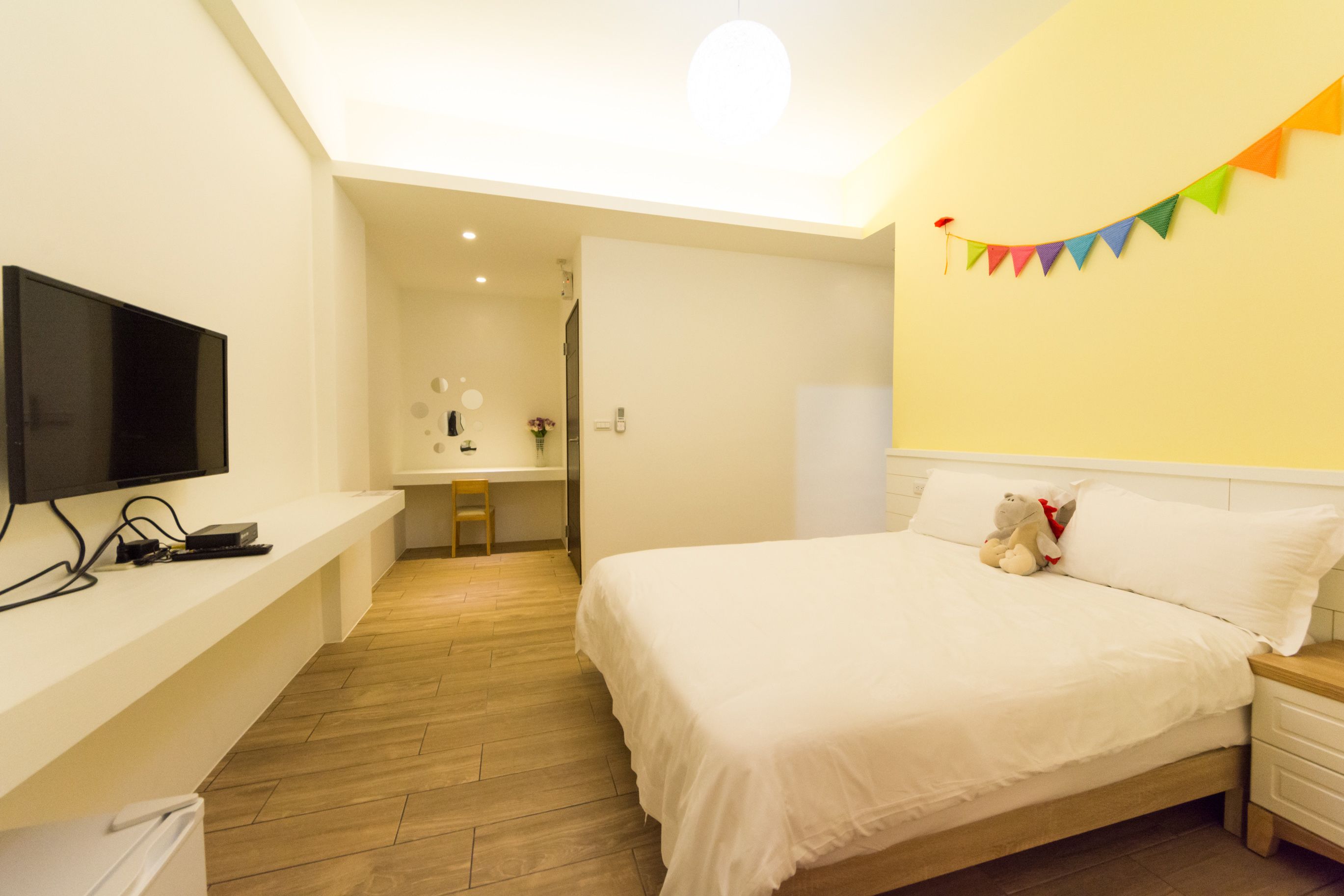 Une chambre avec un lit, un téléviseur posé sur une étagère, un bureau avec une chaise, et une guirlande colorée accrochée au mur.