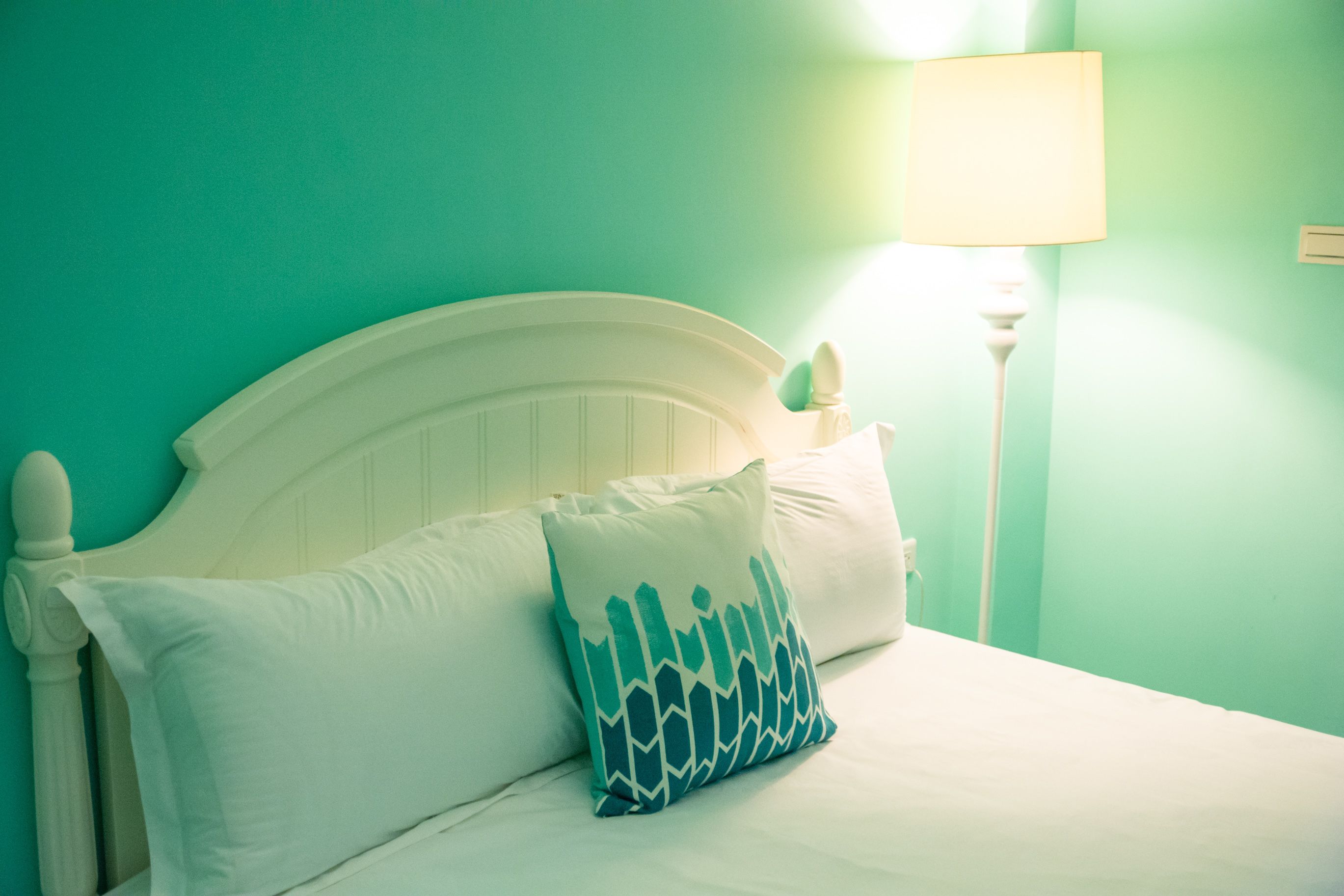 A bed with white bedding and a teal pillow, a white headboard, and a bedside lamp.