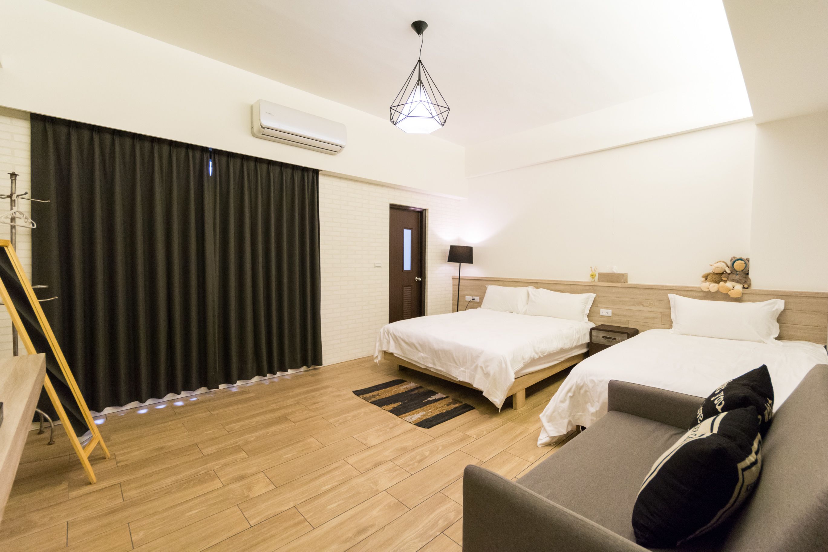 A hotel room with two beds, a sofa, a wooden floor, a black curtain, a lamp, and a hanging light fixture.